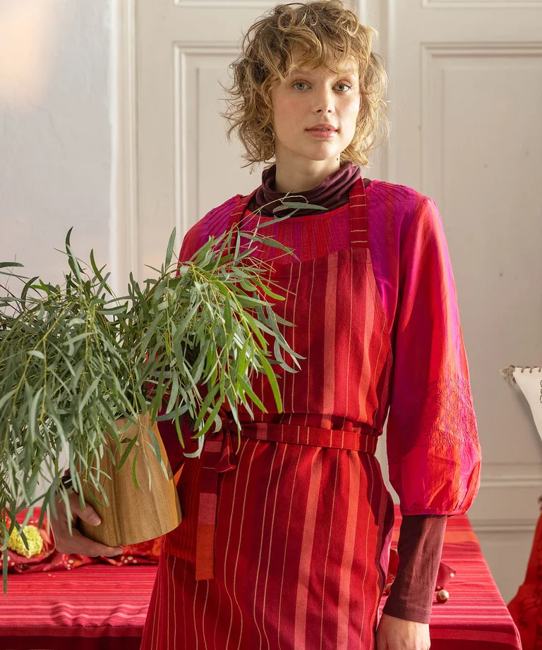A woman holding a potted plant wearing a red, striped apron from Gudrun Sjödén’s 2025 Christmas Home collection.