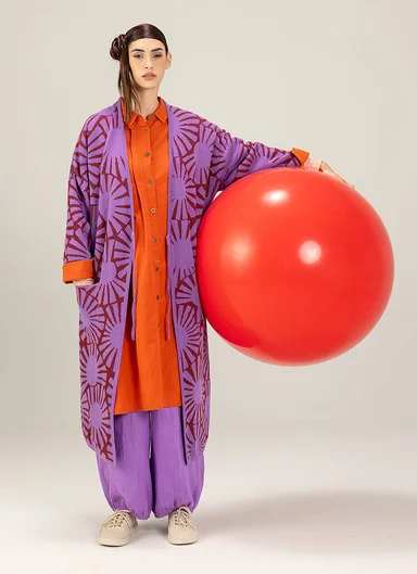 A woman with a large red ball under her arm, wearing layers of purple and red clothing from Gudrun Sjödén.