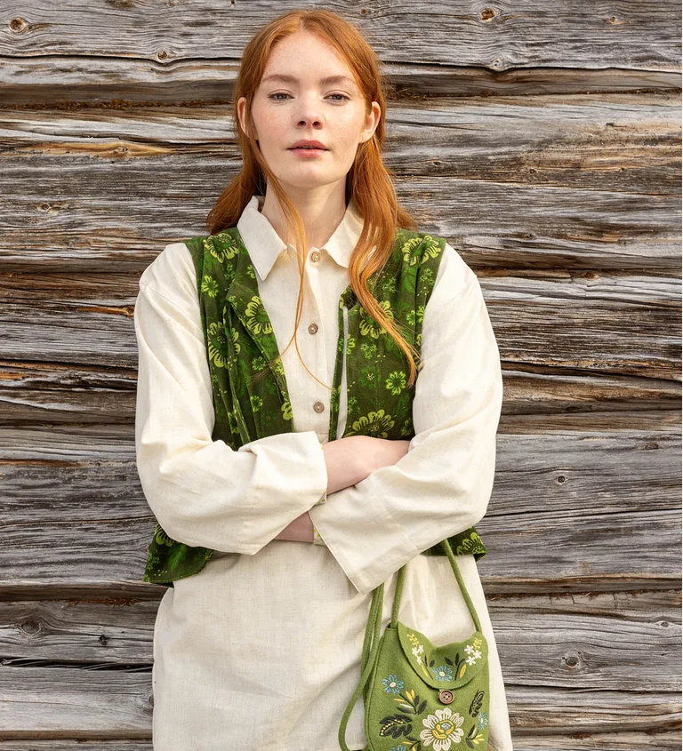 A woman standing with her arms crossed wearing a white shirt and green, patterned waistcoat from Gudrun Sjödén’s 2025 Christmas collection. Holding an embroidered bag in her hand.