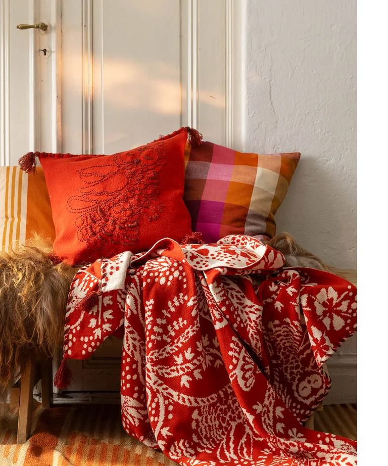 A bench with a red, patterned blanket and two cushions from Gudrun Sjödén’s 2025 Christmas Home collection.