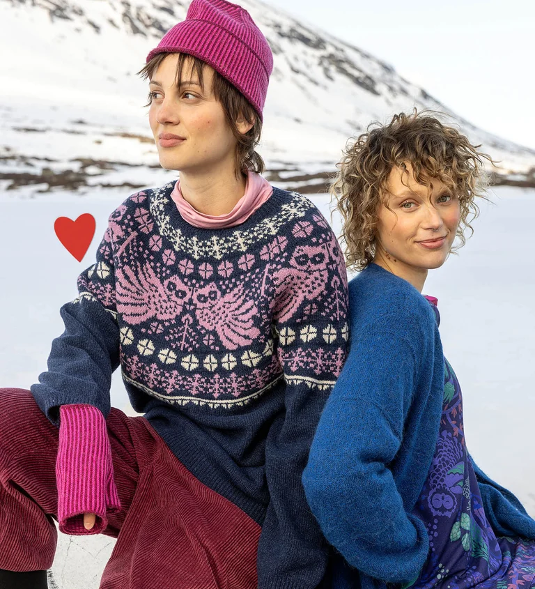 Two women sitting close together wearing clothes from Gudrun Sjödén’s 2025 Christmas collection. There is also a red, graphic heart in the picture.
