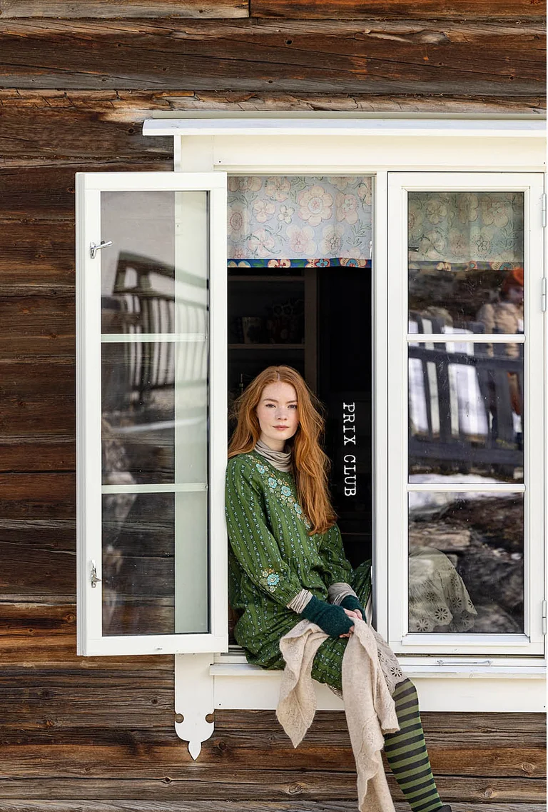 A picture of three women standing next to a timber house wearing clothes from Gudrun Sjödén’s 2025 Christmas collection.