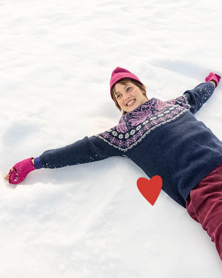 A woman lying down in the snow wearing clothes from Gudrun Sjödén’s 2025 Christmas collection. There is also a red, graphic heart in the picture.