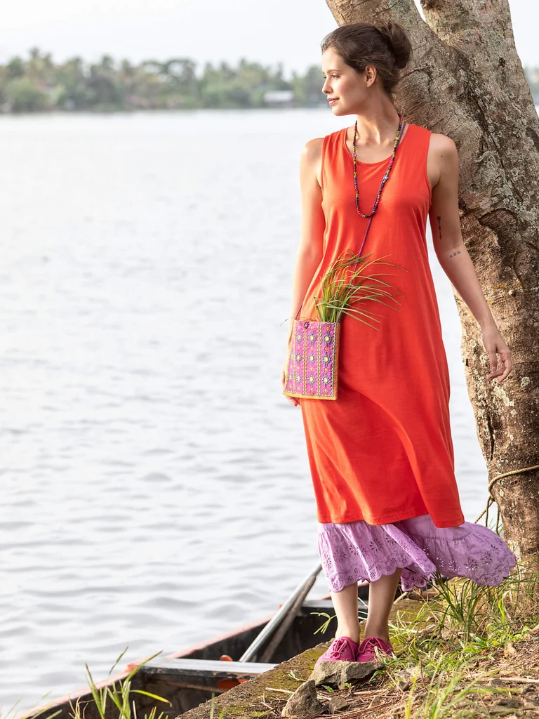 A woman standing by the water wearing a sleeveless orange dress from Gudrun Sjödén.