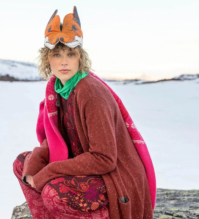 Gudrun Sjödénin vuoden 2025 joulumalliston roosanvärisiin ja punaisiin vaatteisiin kerroksittain pukeutunut nainen, joka istuu kyykyssä eläinnaamio päässä.
