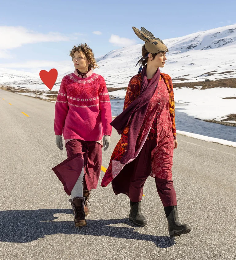 Kaksi Gudrun Sjödénin vuoden 2025 joulumalliston vaatteisiin kerroksittain pukeutunutta naista, jotka kävelevät tietä pitkin. Kuvassa on myös graafinen punainen sydän.