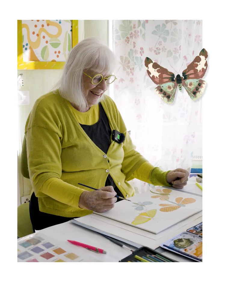 Gudrun Sjödén sitting at a desk painting watercolour butterflies. The picture also includes a standout image of a watercolour butterfly.