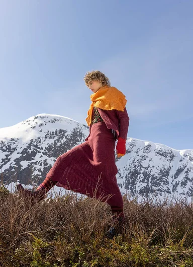 A woman with her arms in the air in a mountain landscape, wearing a burgundy sweater and an orange skirt from Gudrun Sjödén’s Alaska’s Forget-me-not collection.