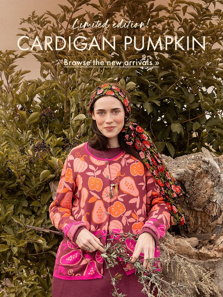 A woman in front of a bush in an autumnal landscape, wearing a patterned knit cardigan from Gudrun Sjödén with orange and pink pumpkins and fruit.