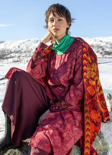 A woman with an animal mask on her head wearing a red and patterned outfit with a knitted cardigan from Gudrun Sjödén’s Alaska’s Wild Heart collection.