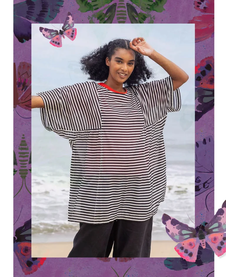 A woman standing on a beach, hands on her head, wearing a purple tunic from Gudrun Sjödén. With a purple frame and standout watercolour butterflies.