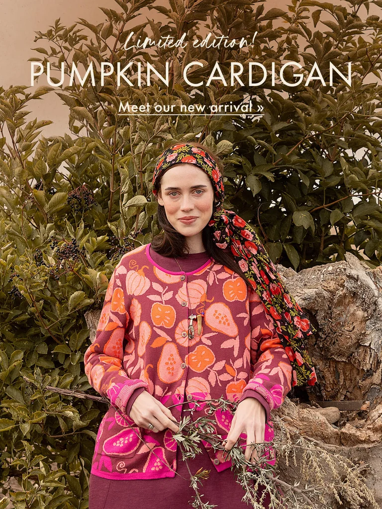 A woman in front of a bush in an autumnal landscape, wearing a patterned knit cardigan from Gudrun Sjödén with orange and pink pumpkins and fruit.