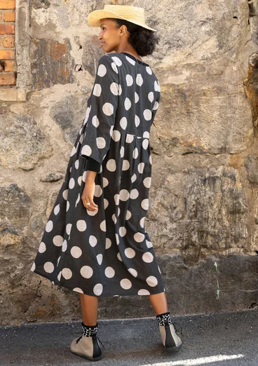 Une femme vêtue d'une robe tissée noire à gros points blancs se tient devant un mur de pierre. On peut voir la vue de dos.