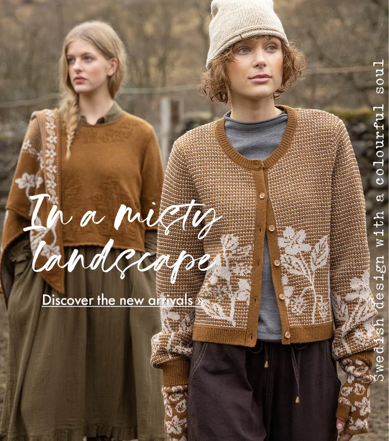 Two women standing next to each other wearing clothes in earthy hues from Gudrun Sjödén. Sheep and a stone wall are visible in the background.