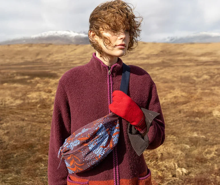 A woman in a mountain landscape wearing a burgundy jacket and red mittens, with a patterned crossbody bag from Gudrun Sjödén over her chest.