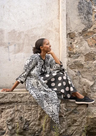 Ein Frau mit einem schwarzem Kleid mit weißem Punkten sitzt auf einer Mauer. Darüber trägt sie ein schwarz-weiß gemustertes Kleid mit Knöpfen welches offen ist.