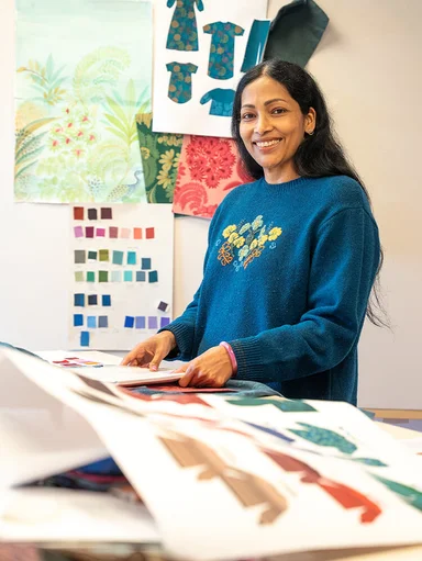 A smiling woman standing by a table with drawings of clothes in the foreground and background at Gudrun Sjödén's headquarters.
