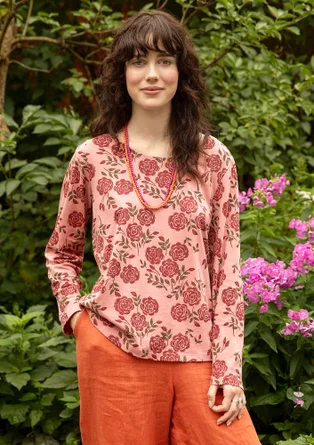 T-shirt rose à manches longues avec des fleurs rouges. Le modèle porte un pantalon orange. 
