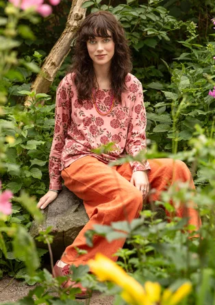 T-shirt rose à manches longues avec des fleurs rouges. Le modèle porte un pantalon orange et est assis sur une pierre entourée de feuilles. 
