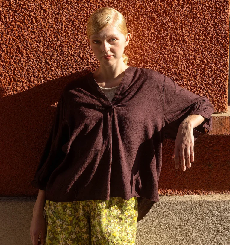 A woman leaning against a wall wearing a brown top and green floral trousers from Gudrun Sjödén.