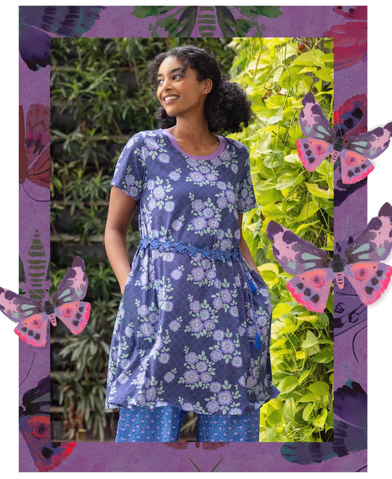 A woman standing on a beach, hands on her head, wearing a purple tunic from Gudrun Sjödén. With a purple frame and standout watercolour butterflies.