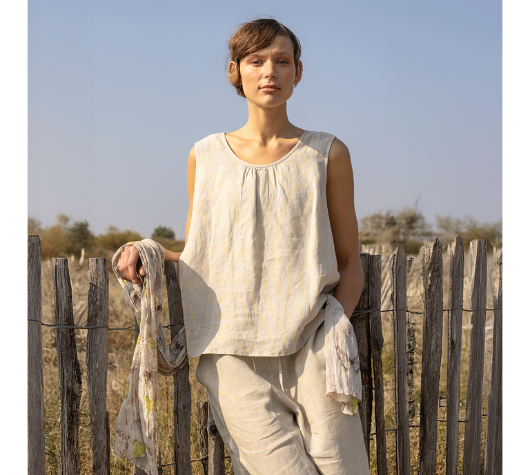 A woman leaning against a fence wearing beige linen clothes from Gudrun Sjödén.