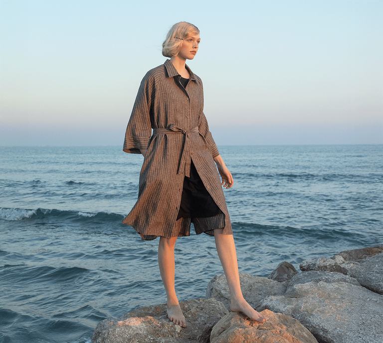 A woman standing on the rocks by the sea wearing a dark grey, striped shirt dress from Gudrun Sjödén.