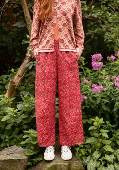 Pantalon rouge de mode naturelle avec un motif de roses rouges et vertes. Avec des chaussures en tissu blanc et un cardigan rose clair avec des boutons et un motif de roses de Gudrun Sjödén.