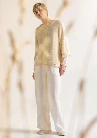 Beige Strickjacke mit Knöpfen. Unter der Strickjacke trägt das Model eine weiss-beiges Streifenshirt. Außerdem hat die Dame eine weisse Hose an. 