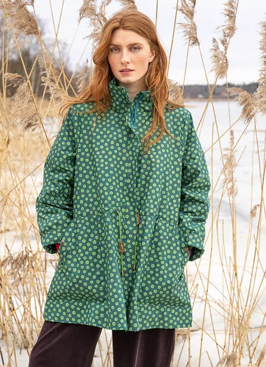 A half picture of a woman wearing a patterned green jacket, with reeds in the background.