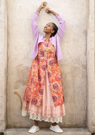 Une femme porte une robe tissée avec des boutons et des bras trois quarts. La robe est orange avec des motifs jaunes, violets et bleus. Sous la robe, elle porte un jupon blanc et un jupon rose clair.  Au-dessus, la femme porte un cardigan ouvert de couleur violet clair. 