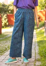 Une femme porte un pantalon bleu et un t-shirt violet de Gudrun Sjödén. Le pantalon a un motif de roses bleu clair et est en tissu de coton bio.