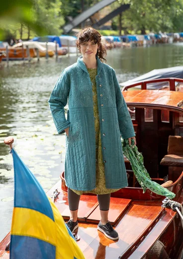 Une femme portant un manteau vert et bleu ouvert se tient sur un bateau en bois. Le manteau est orné de broderies claires. La femme porte également une robe verte, un leggins sombre et des chaussures en tissu sombre et tient une écharpe verte dans sa main gauche.