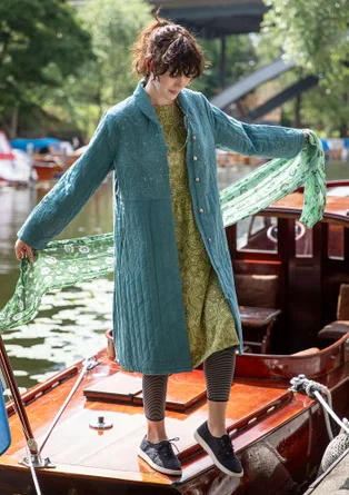 Une femme portant un manteau vert et bleu ouvert se tient sur un bateau en bois. Le manteau est orné de broderies claires. La femme porte également une robe verte, un leggins sombre et des chaussures en tissu sombre et tient une écharpe verte dans sa main gauche.