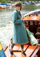 Une femme portant un manteau vert et bleu ouvert se tient sur un bateau en bois. Le manteau est orné de broderies claires. La femme porte également une robe, un leggins et des chaussures en tissu sombre.