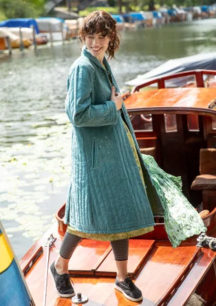 Une femme portant un manteau vert et bleu ouvert se tient sur un bateau en bois. Le manteau est orné de broderies claires. La femme porte également une robe, un leggins et des chaussures en tissu sombre.