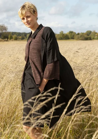 Die Dame trägt eine braune Strickjacke mit dreiviertel Armen und Knöpfen. Darunter trägt sie eine schwarze Bluse und eine schwarze, kurze Hose. Über der Strickjacke hat das Model noch ein schwarzes Kleid an.