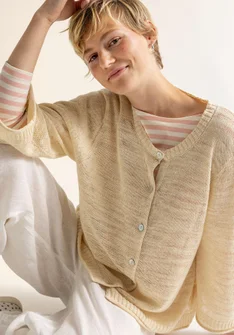 Model trägt eine beige Strickjacke mit Knöpfen. Unter der Strickjacke trägt die Frau eine weiss-beiges Streifenshirt. Außerdem hat die Dame eine weisse Hose an. 