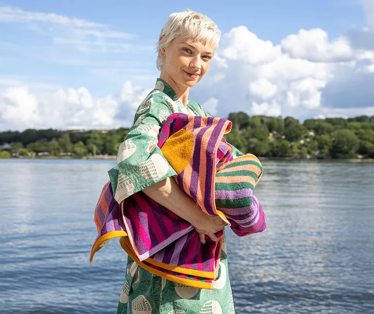 A woman by a lake holding striped terry towels from Gudrun Sjödén.