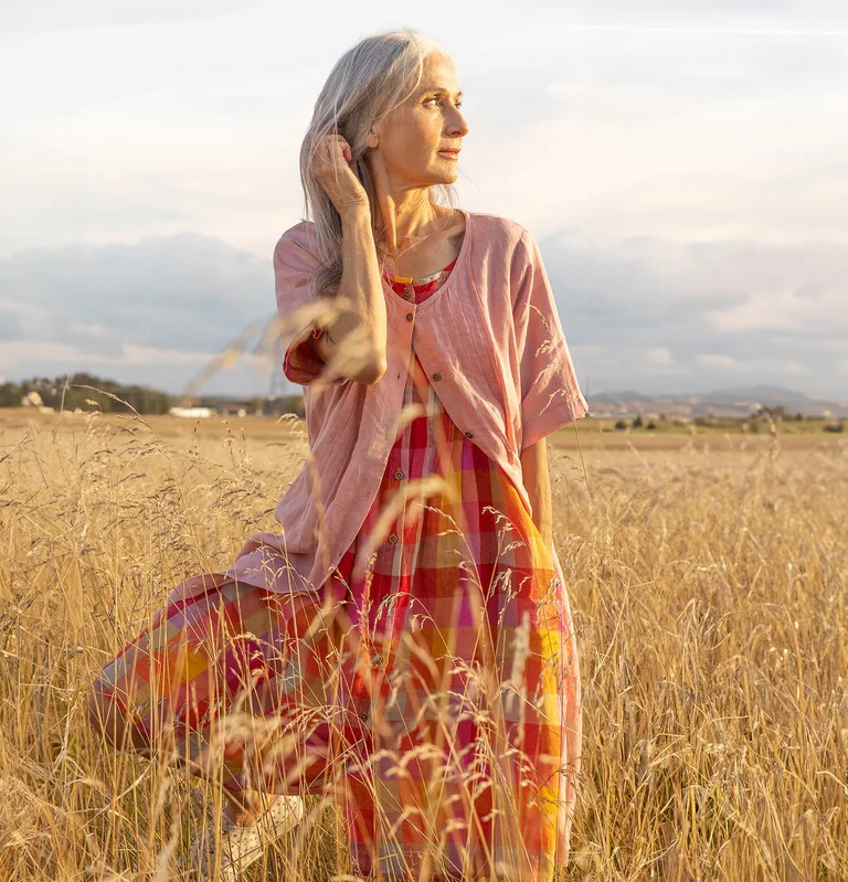 A woman in a flax field wearing a pink check dress and pink cardigan from Gudrun Sjödén 