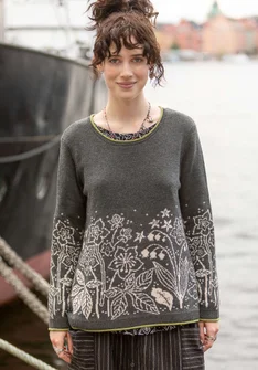 Une femme se tient sur le ponton d'un bateau, vêtue d'un pull gris foncé. Un motif floral clair est brodé sur le bas du haut.