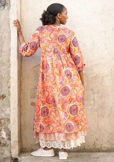 Eine Frau trägt ein Webkleid mit Knöpfen und dreiviertel Armen. Das Kleid ist orange mit Mustern in gelb, violett und blau. Unter dem Kleid trägt sie einen weißen und einen hellroséfarbenen Unterrock.  Es ist die Rückenansicht zu sehen. 