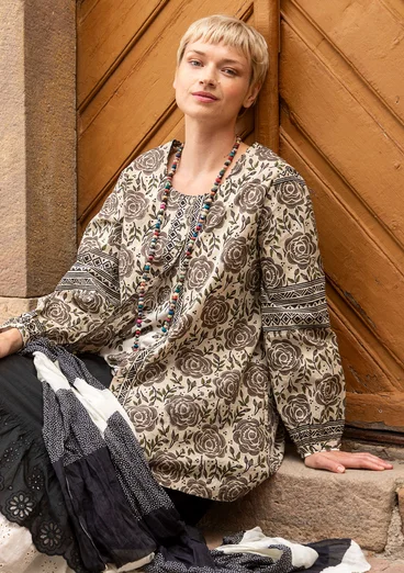 La femme porte des vêtements naturels de Gudrun Sjödén - une tunique en coton beige avec des boutons et un motif de roses marron, avec un jupon noir.