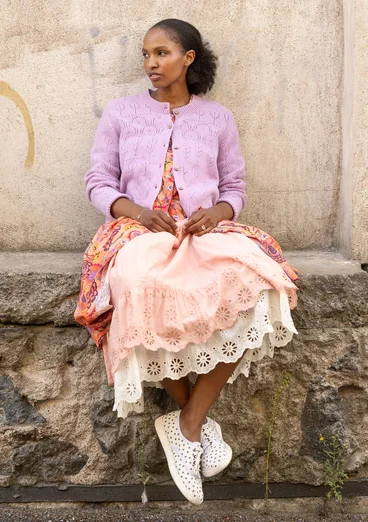 Cardigan à manches longues avec technique de tricotage pointelle dans la couleur lilas. Combiné avec une robe orange avec un motif floral coloré et deux jupes avec de la dentelle dans les couleurs blanc et rose. Le modèle porte des chaussures blanches à pois noirs. 
