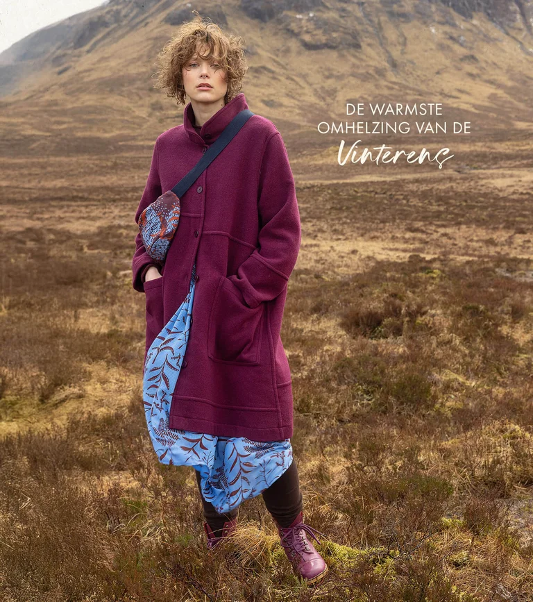 A woman in a mountain landscape wearing a burgundy wool coat, a light blue patterned dress and a crossbody bag from Gudrun Sjödén.