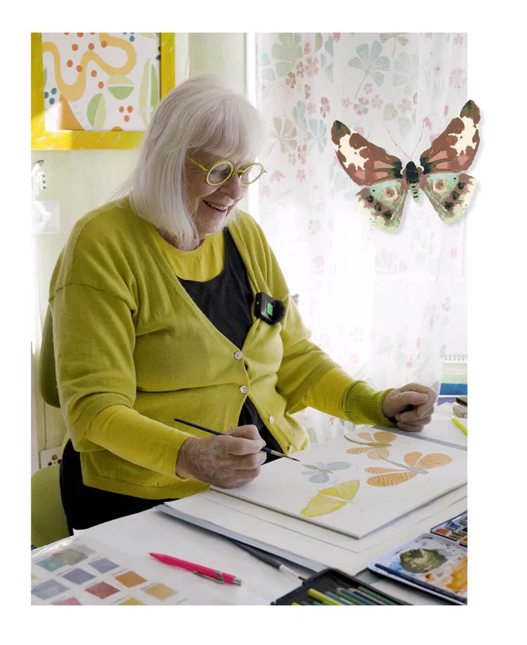 Gudrun Sjödén sitting at a desk painting watercolour butterflies. The picture also includes a standout image of a watercolour butterfly.