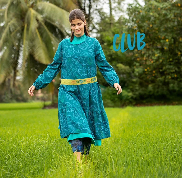 A woman walking on a green lawn wearing a blue, patterned dress and a lime-green fabric belt from Gudrun Sjödén.