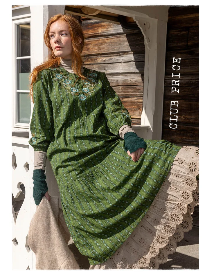 A woman sitting on a railing wearing layers of green and beige clothes from Gudrun Sjödén’s 2025 Christmas collection.