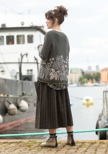 Eine Dame steht am Bootssteg mit dunkelgrauem Pullover. Der untere Teil des Oberteils ein helles Blumenmuster aufgestickt. Untenrum trägt die Dame einen Rock in schwarz und mit Streifen.