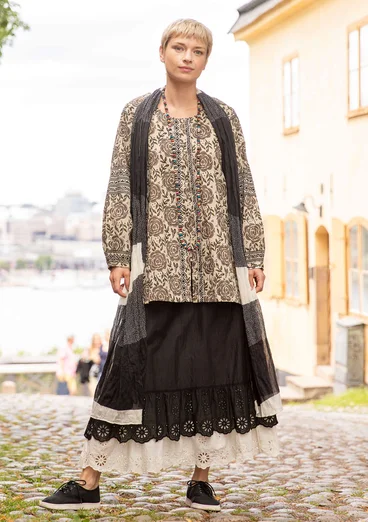 La femme porte des vêtements naturels de Gudrun Sjödén - une tunique en coton beige avec des boutons et un motif de roses marron. Elle l'accompagne d'un jupon noir, d'un foulard noir et de chaussures en tissu noir.